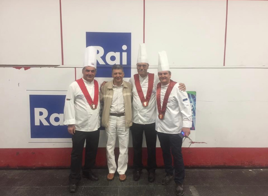 Four people standing together in chef jackets and medals against a wall with blue-and-white signage, evenly lit.