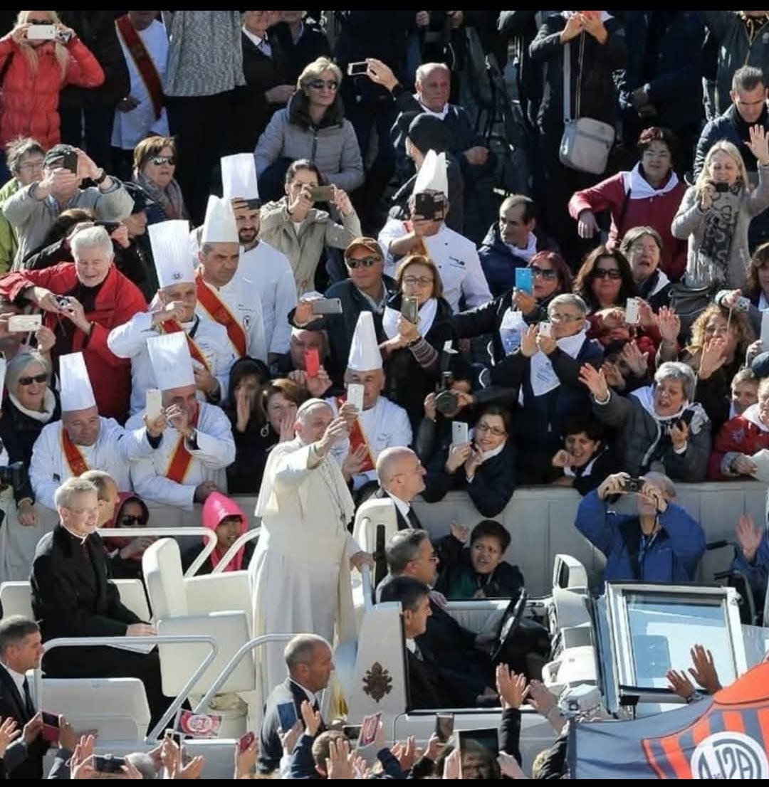 A large outdoor crowd surrounding several people in white chef hats and ceremonial clothing, with many spectators holding up phones in bright daylight.