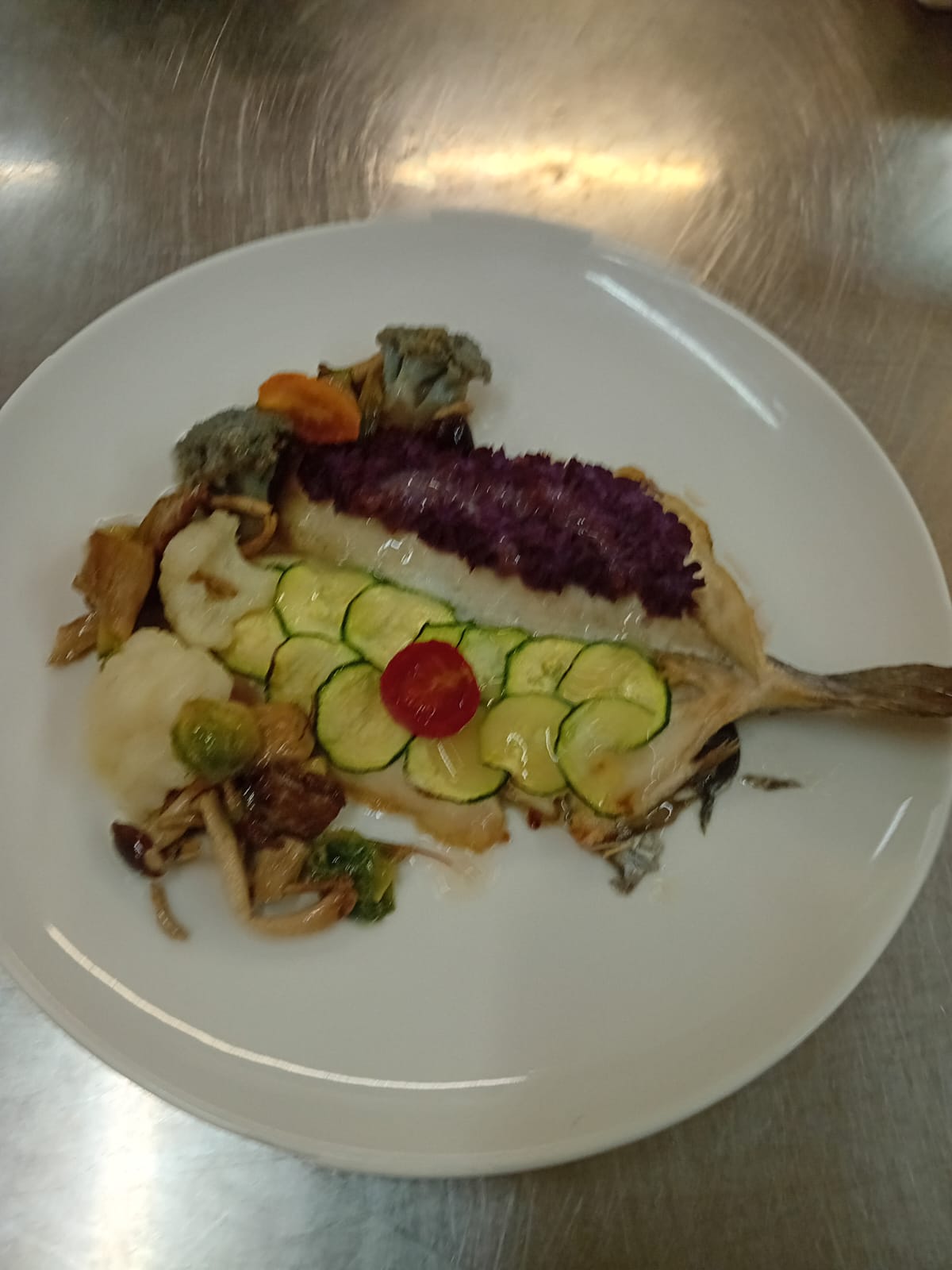 A plated fish dish topped with sliced zucchini, purple garnish and vegetables on a white plate, under overhead lighting.