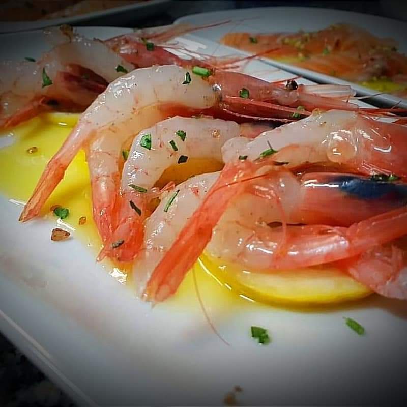 Close-up of prawns arranged on a white plate with oil and herbs, softly lit.