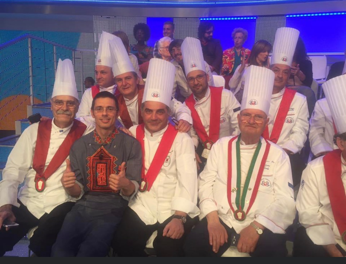 Group of people in chef hats and ceremonial sashes pose closely together on a blue-lit stage.