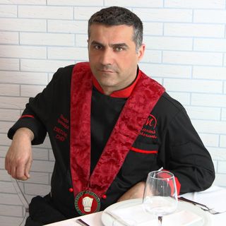 A seated man in a black outfit with a red sash at a table, framed from the waist up against a light brick wall.