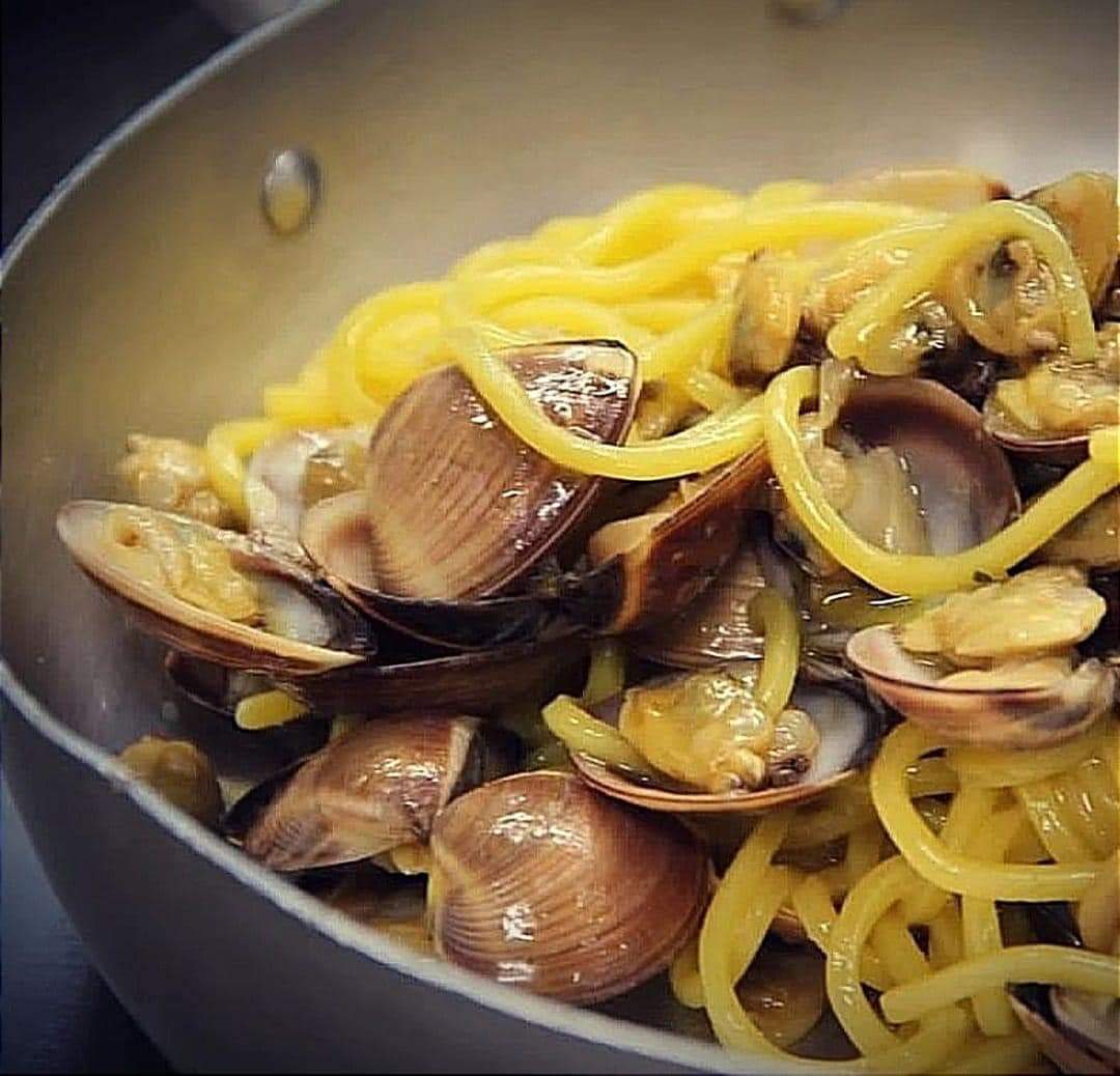 Close-up of pasta with open clams in a pan, warmly lit.