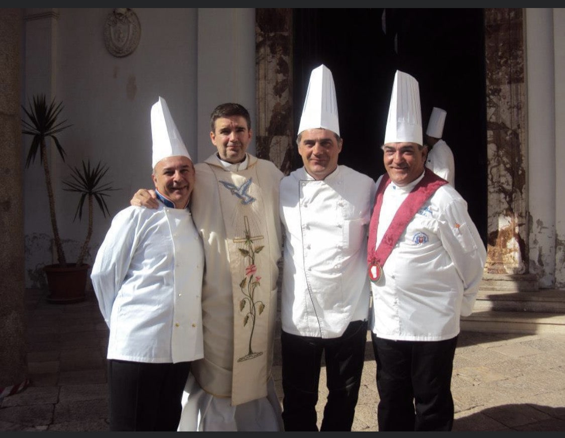 Four people in chef attire and ceremonial clothing pose outdoors in bright sunlight.