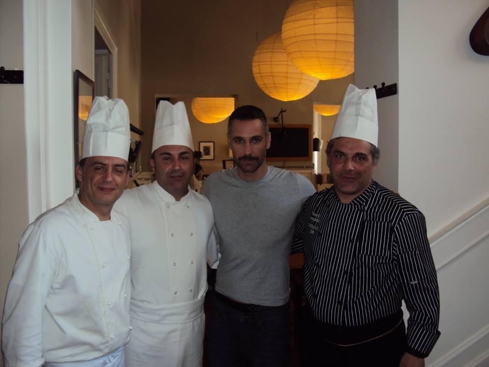 Four people, including three chefs, pose indoors beneath large warm pendant lights.