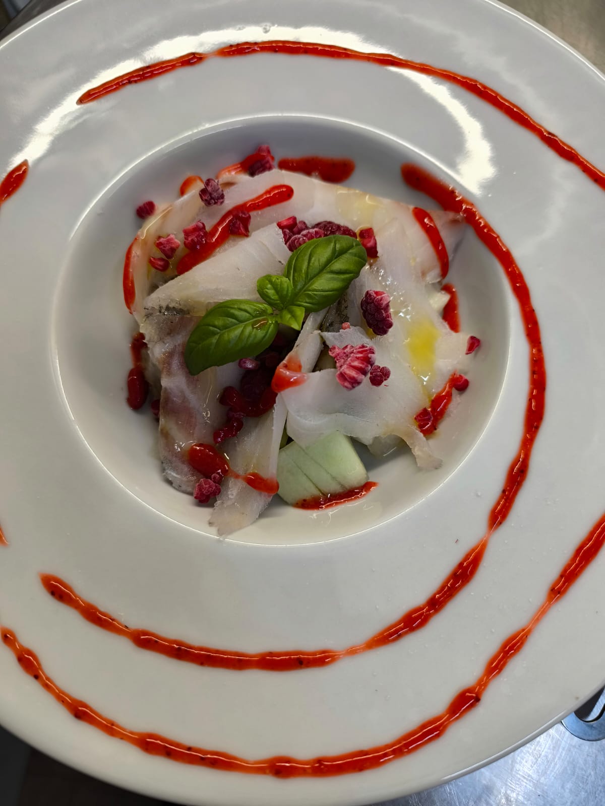 A plated dish of pale sliced pieces with basil, red sauce and berry garnish, viewed from above under bright lighting.
